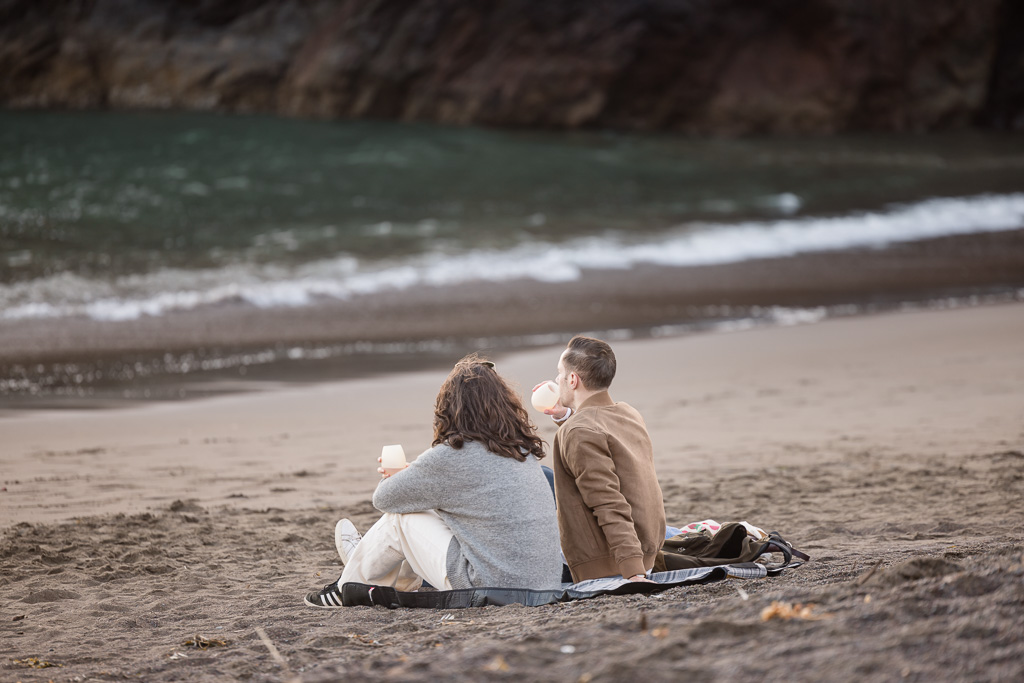 couple sitting on a picnic blanket on the beach enjoying a drink